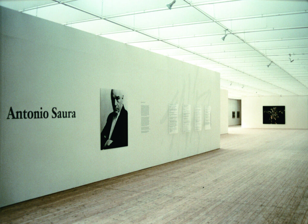 Bild över Antonio Sauras utställning. En vit vägg med text samt ett svartvitt porträtt av Antonio.
