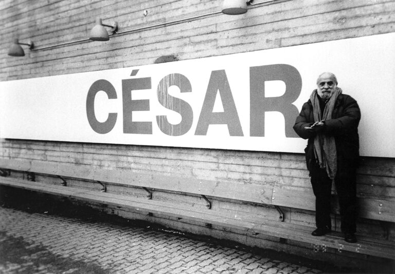 En stor skylt med texten "César" i versaler pryder väggen utanför ingången till konsthallen. Under skylten är en bänk och till höger i bild står César själv på bänken bredvid skylten.