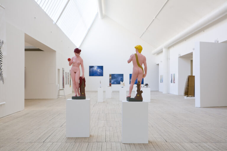 Färglada skulpturer av naken man och kvinna står på podier i konsthallens ljusschakt.