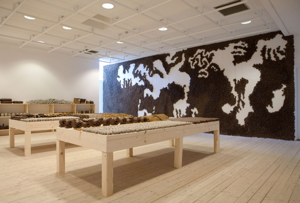 Översiktsbild av ett rum i utställningen Anna Maria Maiolino, 2011. Installationer i lera på väggen och på bord runtom i lokalen.