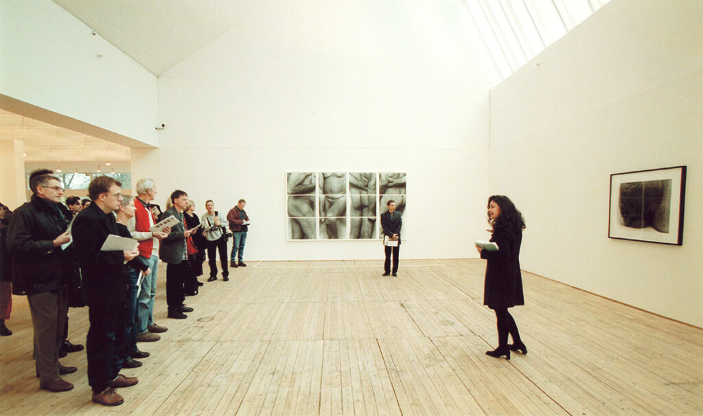 En inlyssnande publik på visning av John Coplans fotografier. Besökarna står vända mot en talare som berättar om verken.