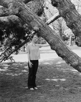 Svartvit bild av kvinna som står under en tall och håller sin hand på trädets stam.