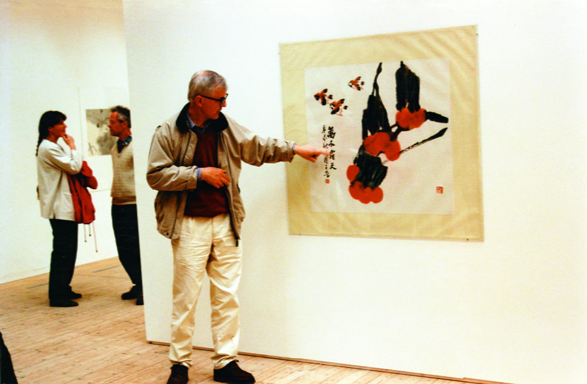 A man stands in front of a drawing in the art gallery and points to a detail in the picture. The man has glasses and is wearing a beige jacket and white trousers.