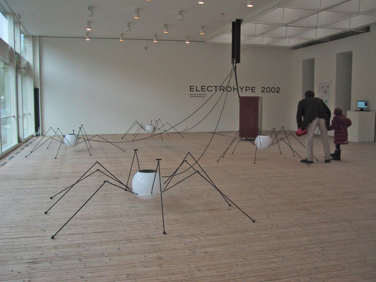 Installationsbild från utställningen Electrohype 2002. Ett par besökare betraktar spindellika skulpturer som står utspridda på trägolvet.
