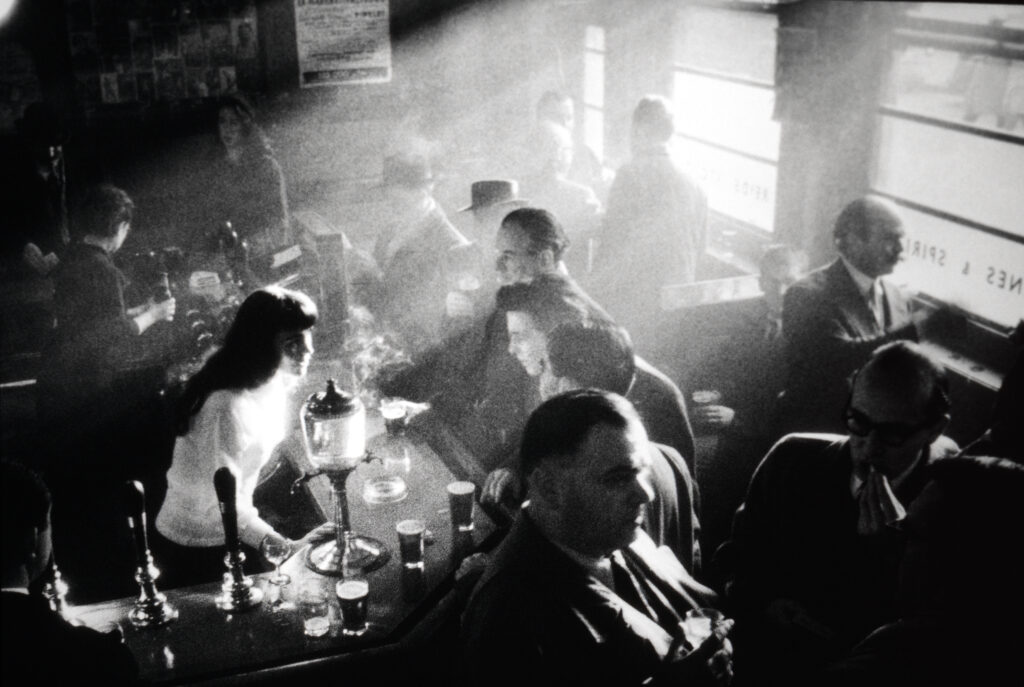 Svartvit bild av människor som umgås och dricker i en fullsatt bar, och en bartender som serverar drinkar. Solljus strömmar in genom fönstren.