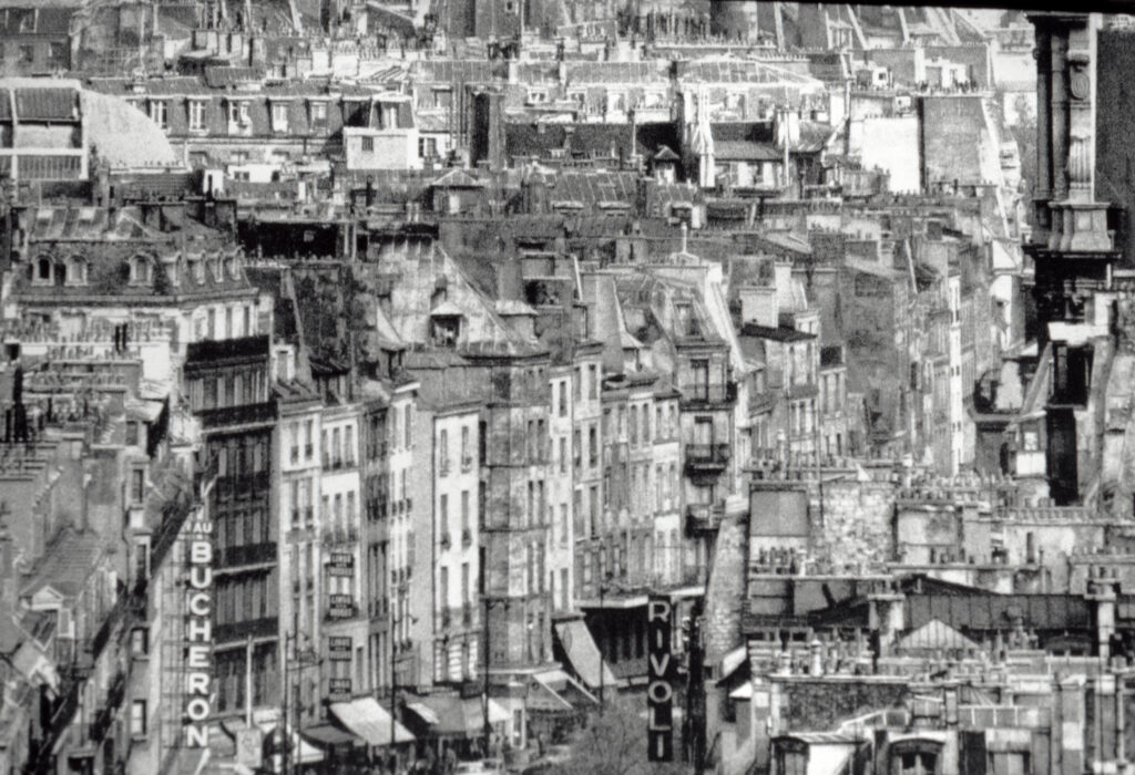Svartvit bild av en tät stadsbebyggelse med äldre husfasader, skyltar och taklandskap