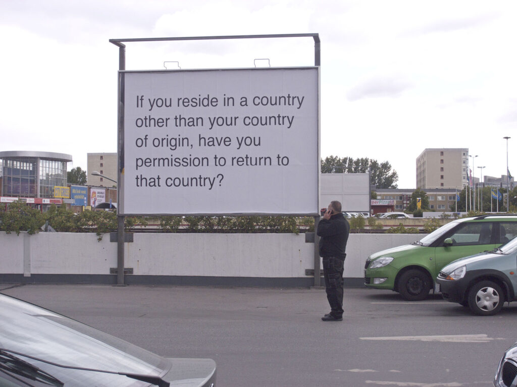 En stor billbord-tavla med texten "If you reside in a country of origin, have you permission to return to that country? Nedanför står en man med och tittar på texten med en telefon.