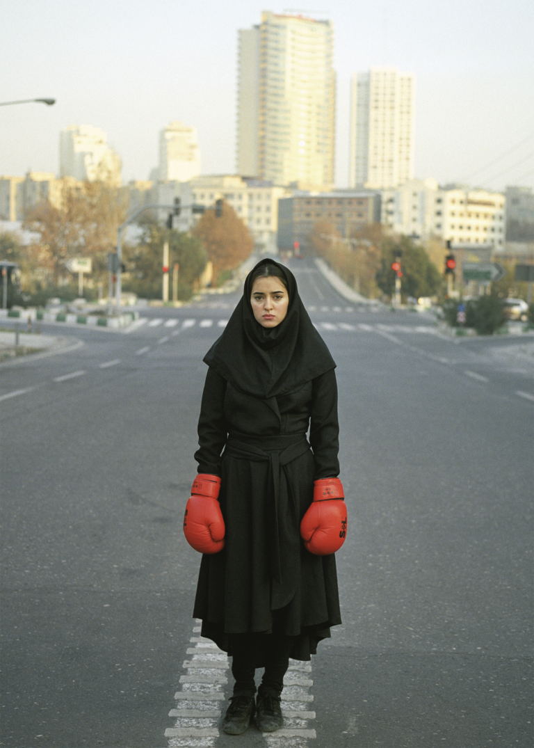 Foto av ung kvinna kädd i svart dräkt, hijab och röda boxningshandskar. Hon står mitt i en väg.