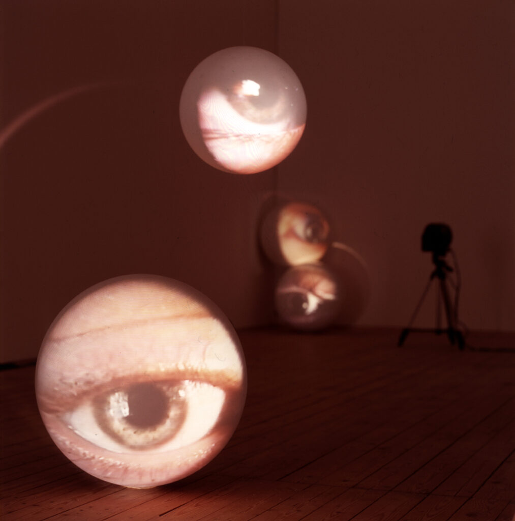 Mänskliga ögon projiseras på ljusa bollar i olika storlekar. Den närmsta bollen ligger på golvet, och en annan är svävande. I bakgrunden syns två bollar till med ögon, som står lutade mot väggar i ett hörn ovanpå varandra.