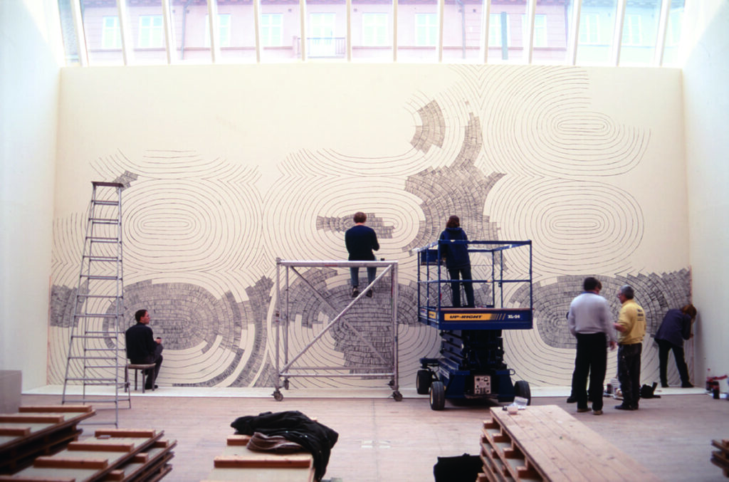 Bild på uppbyggnaden av Olav Christopher Jenssens utställning. Flera personer står vid väggen och målar, en person står i en skylift och en sitter på en byggställning.
