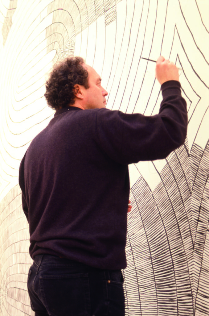 Bild av Olav Christopher Jenssen när han målar sitt verk på en av konsthallens väggar. Bilden är tagen snett underifrån. Olav är klädd i svart och har mörkt lockigt hår.