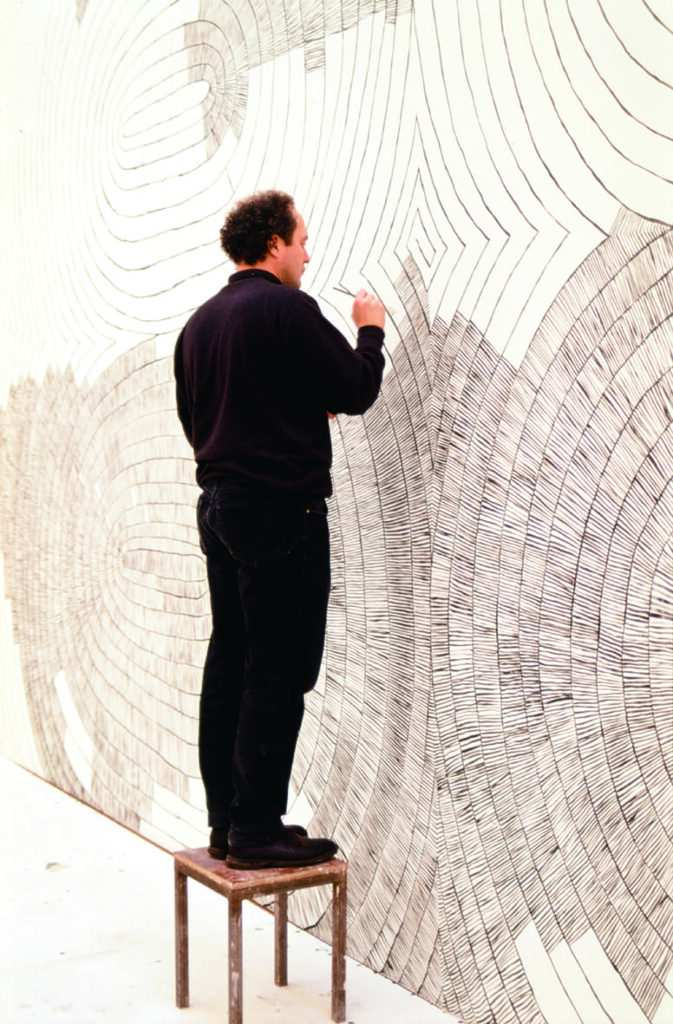 Bild av Olav Christopher Jenssen när han målar sitt verk på en av konsthallens väggar. Olav är klädd i svart och har mörkt lockigt hår, han står på en pall med en pensel i ena handen.