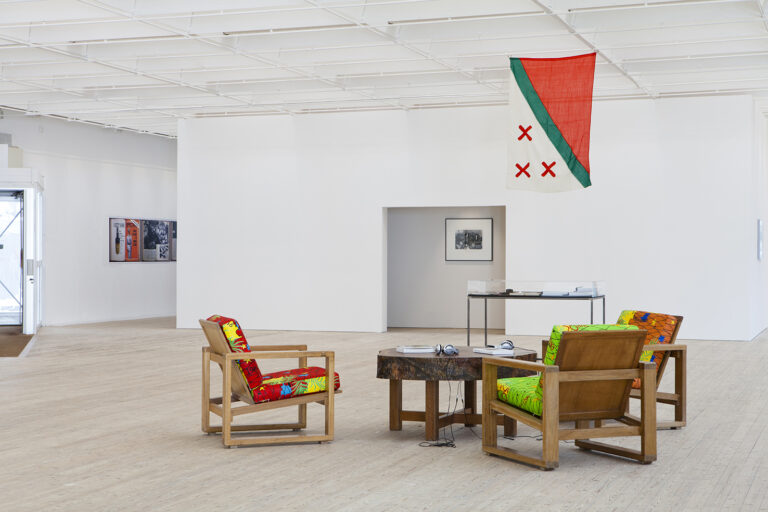 Grova trästolar med kuddar i glada färger står runt ett bord i konsthallen. En flagga i rött, grönt och vitt hänger över bordet.