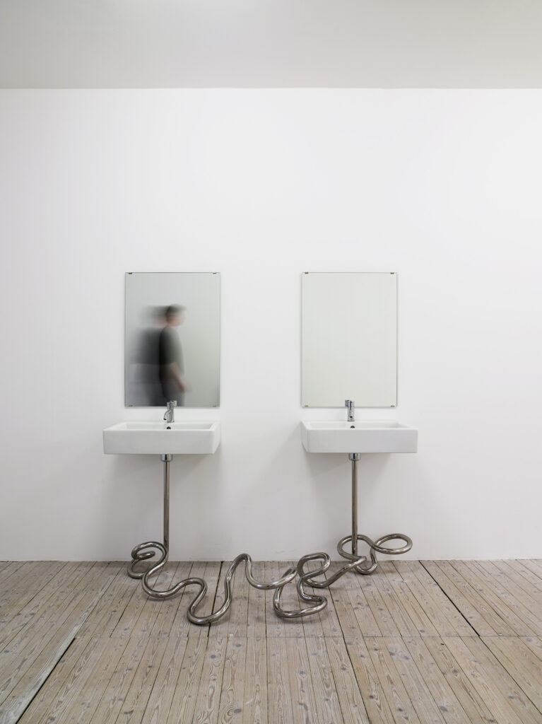 Installation av Elmgreen och Dragset. Två handfat med speglar sitter bredvid varandra. Avloppen under vaskarna sitter ihop och i en organiskt slingrande form.