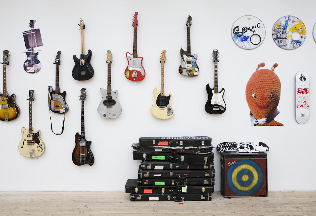 Elgitarrer och basgitarrer hänger på en vägg, under ligger deras fordral, en skateboard och en högtalare.