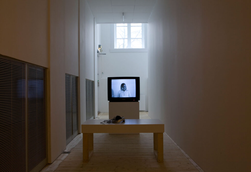 Installationsbild/vy av utställningen Throwing Three Balls in the Air, 2010. En monitor/TV med en person i bild och en liten bänk att sitta på.