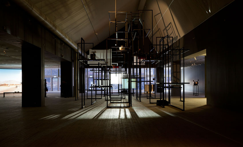 En stor metallkonstruktion sträcker sig upp i Malmö Konsthalls mörklagda ljusschakt. I bakgrunden anar man filmprojektioner.