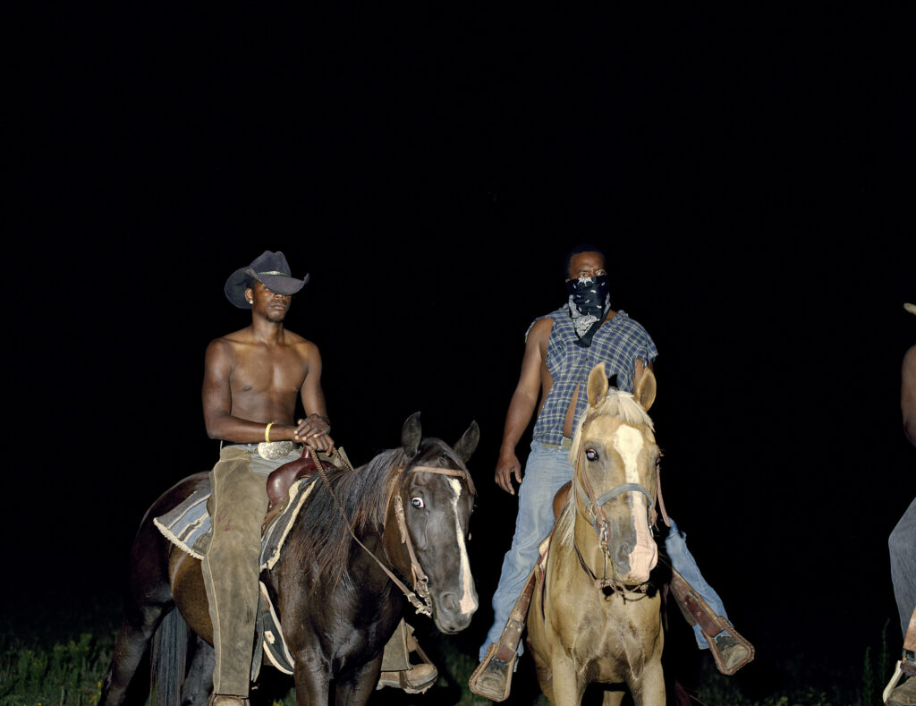 Two men in relaxed cowboy style ride on separate horses. The picture is taken in darkness with strong flash.