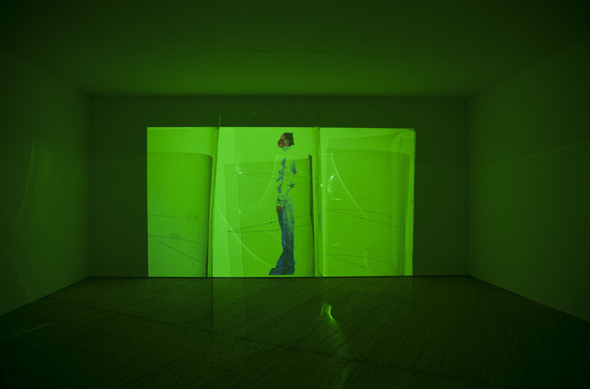 Fotografi av film som spelar på väggen i ett mörklagt rum. Filmen lyser i neongrönt, en kvinna syns i bild.