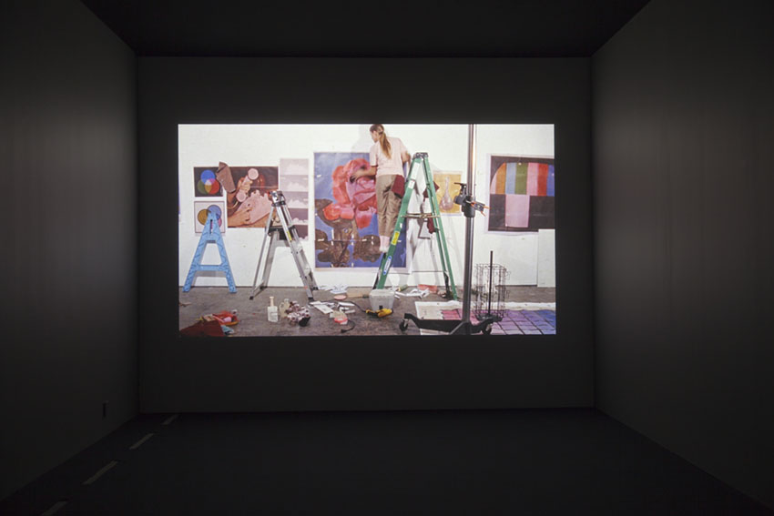 Bild av film på väggen i ett mörkt rum. En person står på en stege och monterar en färglad ros på väggen.