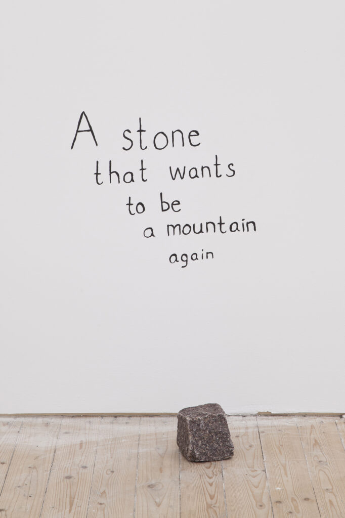 A cobblestone is on the floor in front of the text. "A stone that wants to be a mountain again".