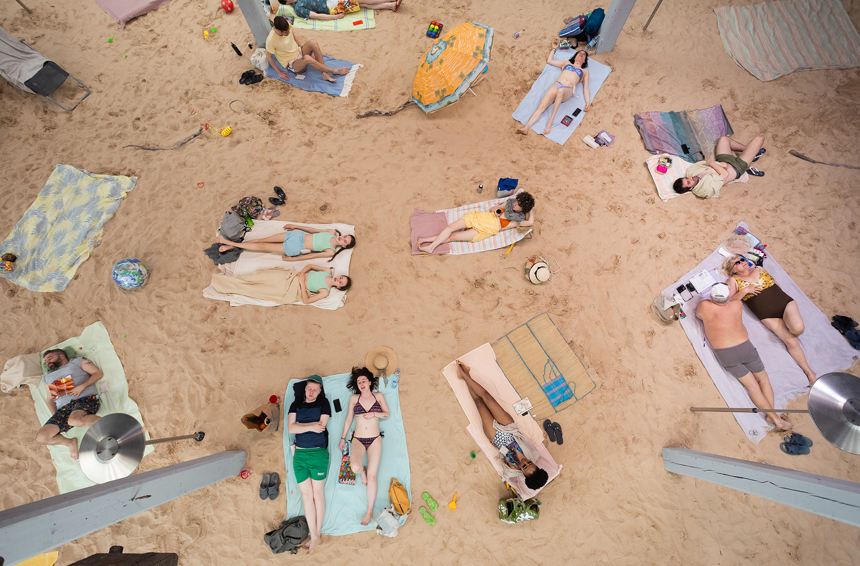 Top-down photo of people lying on a sandy beach, singing, wearing pastel-colored swimwear.