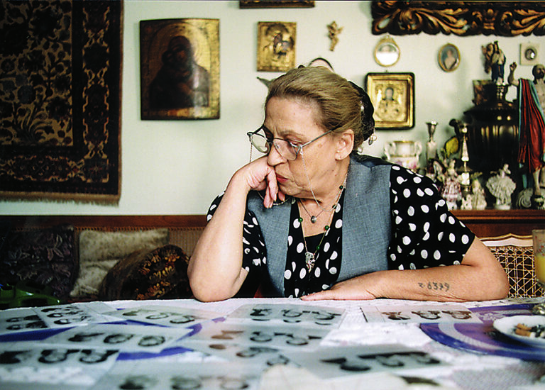 Ceija Stojka sitter vid ett bord och tittar på porträttfotografier. I bakgrunden ser man en vägg med olika ikoner och helgon.