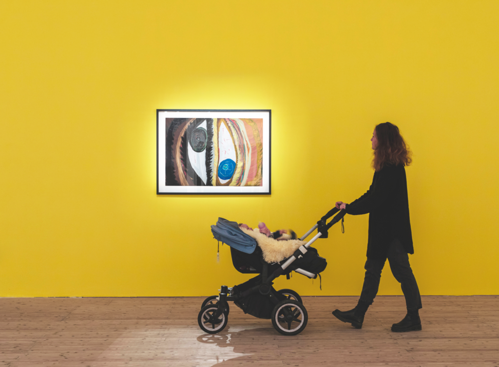 A mother with a stroller in front of a yellow wall in the exhibition. On the wall, there is a piece featuring two eyes: one blue and one brown.