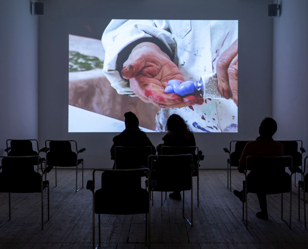 The film room in the exhibition where a documentary about Ceija Stojka's life is being screened. Three people are sitting in the darkness in front of the film.