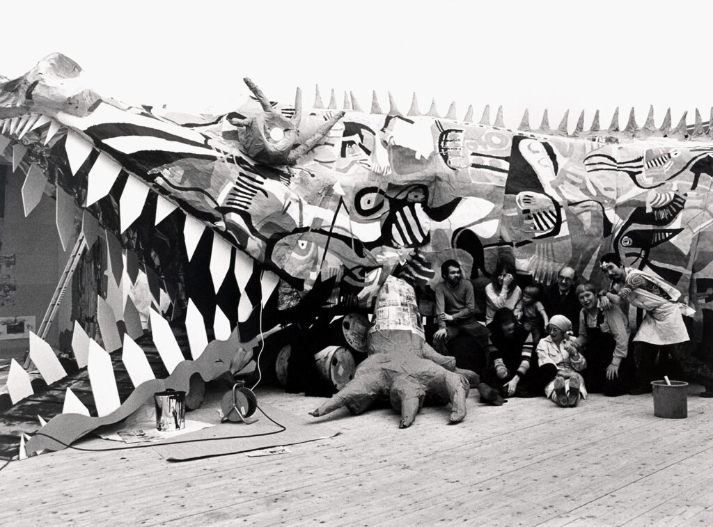 Black and white image of a large crocodile sculpture with people posing in front of the sculpture. 