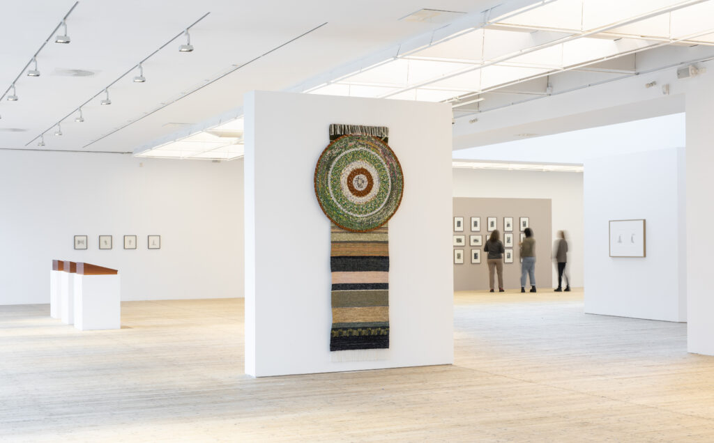 Exhibition view depicting a large crocheted piece hanging on the wall, shaped like a rug with a circle on top. In the background, visitors can be seen in the bright art gallery building.