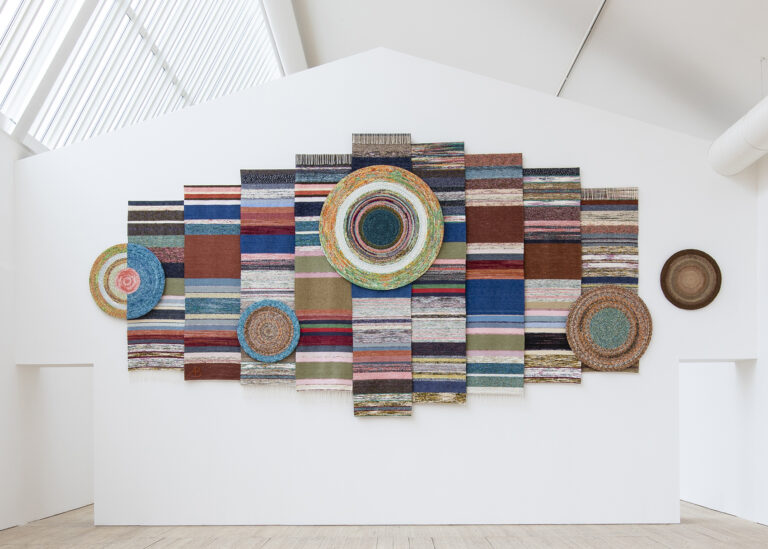 I ljusschaktet i konsthallens utställningsrum hänger Ann Böttchers färgrika verk gjort av mattrasor på en vit vägg.
