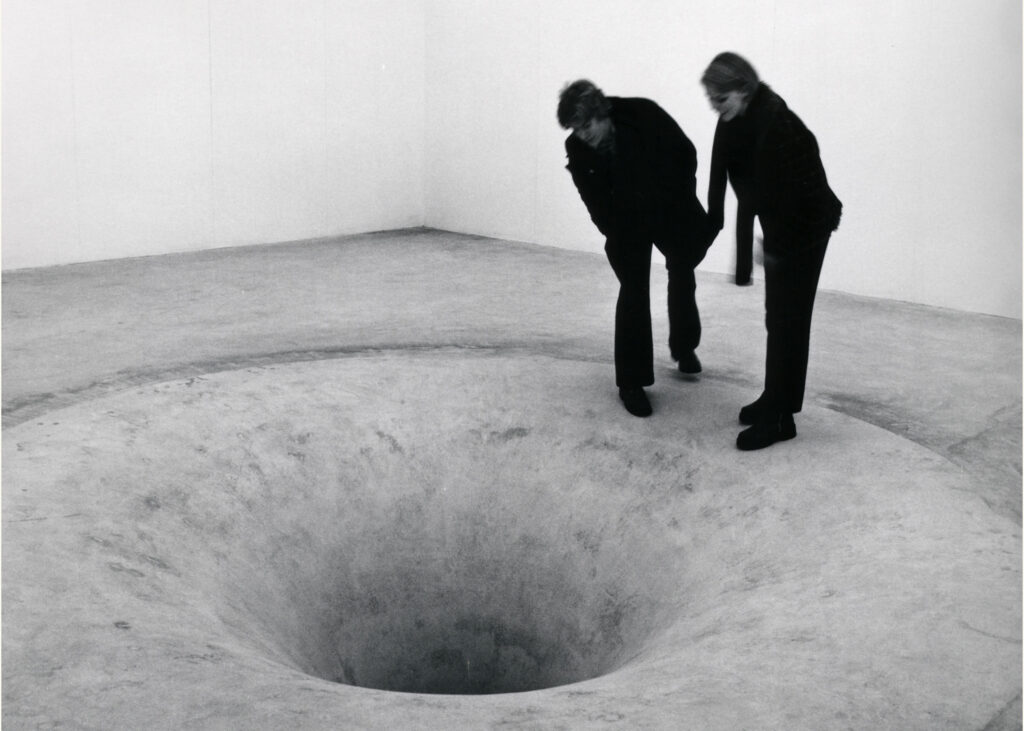 Black and white image of people peering down into what appears to be a bottomless concrete hole in the art gallery's floor.