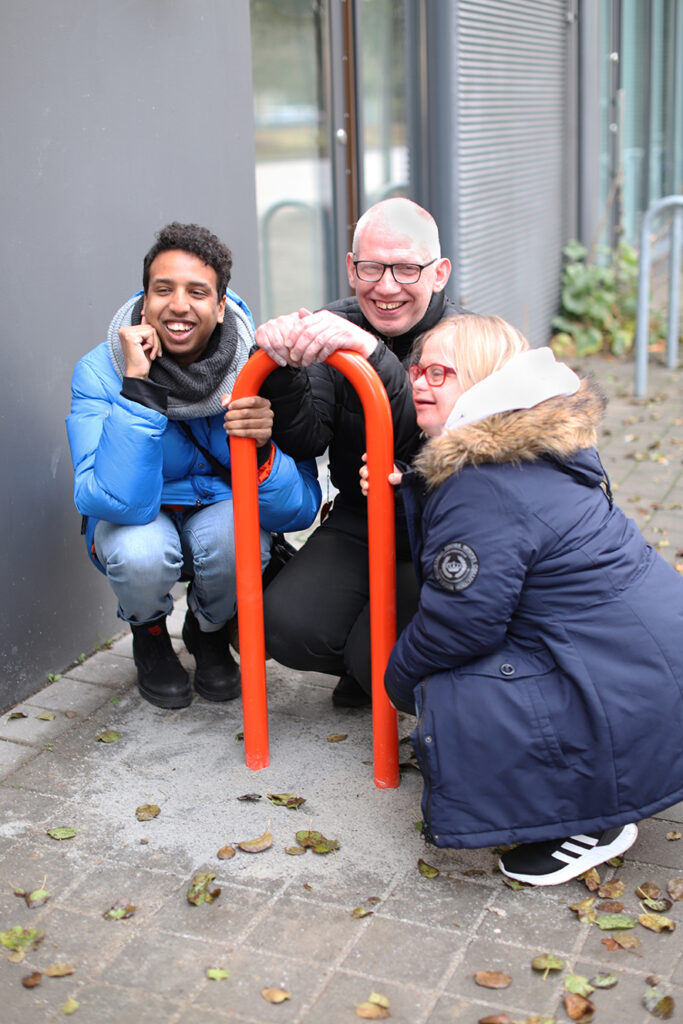 Three people are squatting on the ground by a red bicycle rack and smiling.