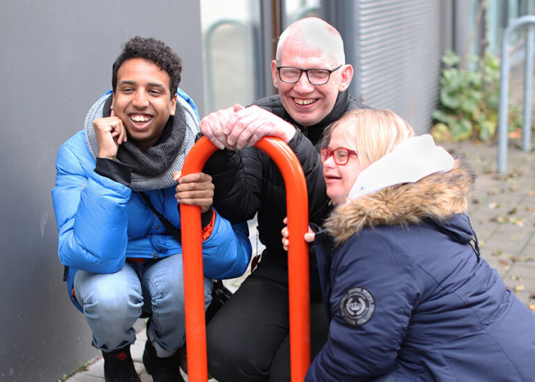 Three people are smiling and squatting by a red bicycle rack