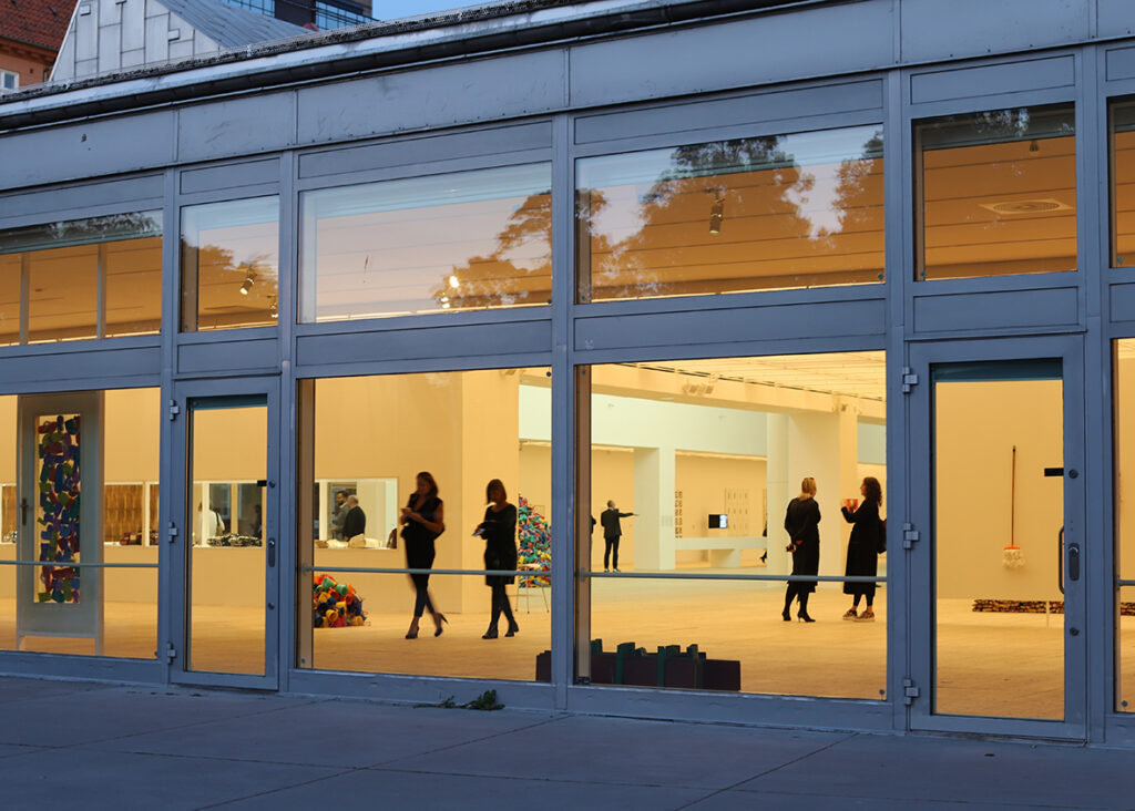 Bild tagen i skymningen in genom konsthallens fönster. Personer rör sig inne i det upplysta rummet.
