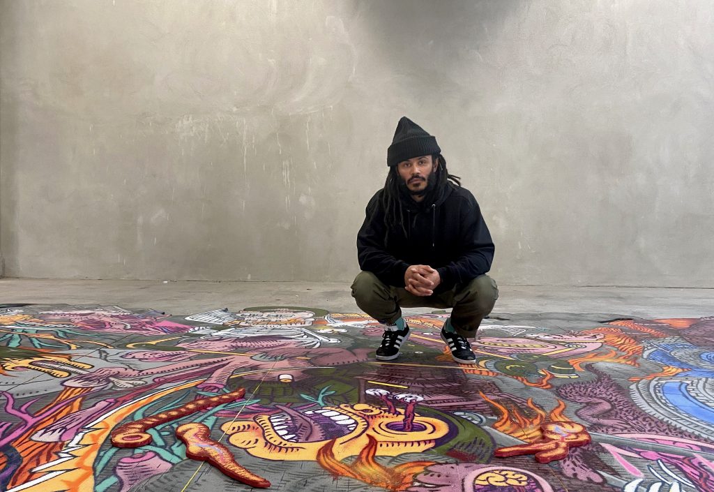 Image of artist Kim Demåne crouching next to one of their graffiti works.