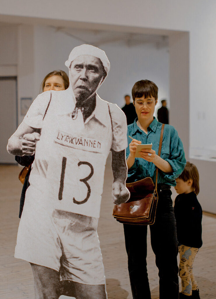 Bild av besökare i utställningshallen, två kvinnor och ett barn bakom Åke Hodell som pappfigur.