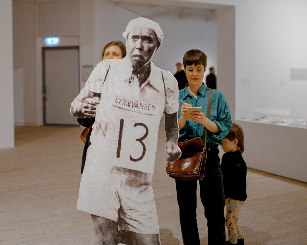 Visitors in the exhibition hall, two females and one child are standing behind Åke Hodell as a large paper cut out. 