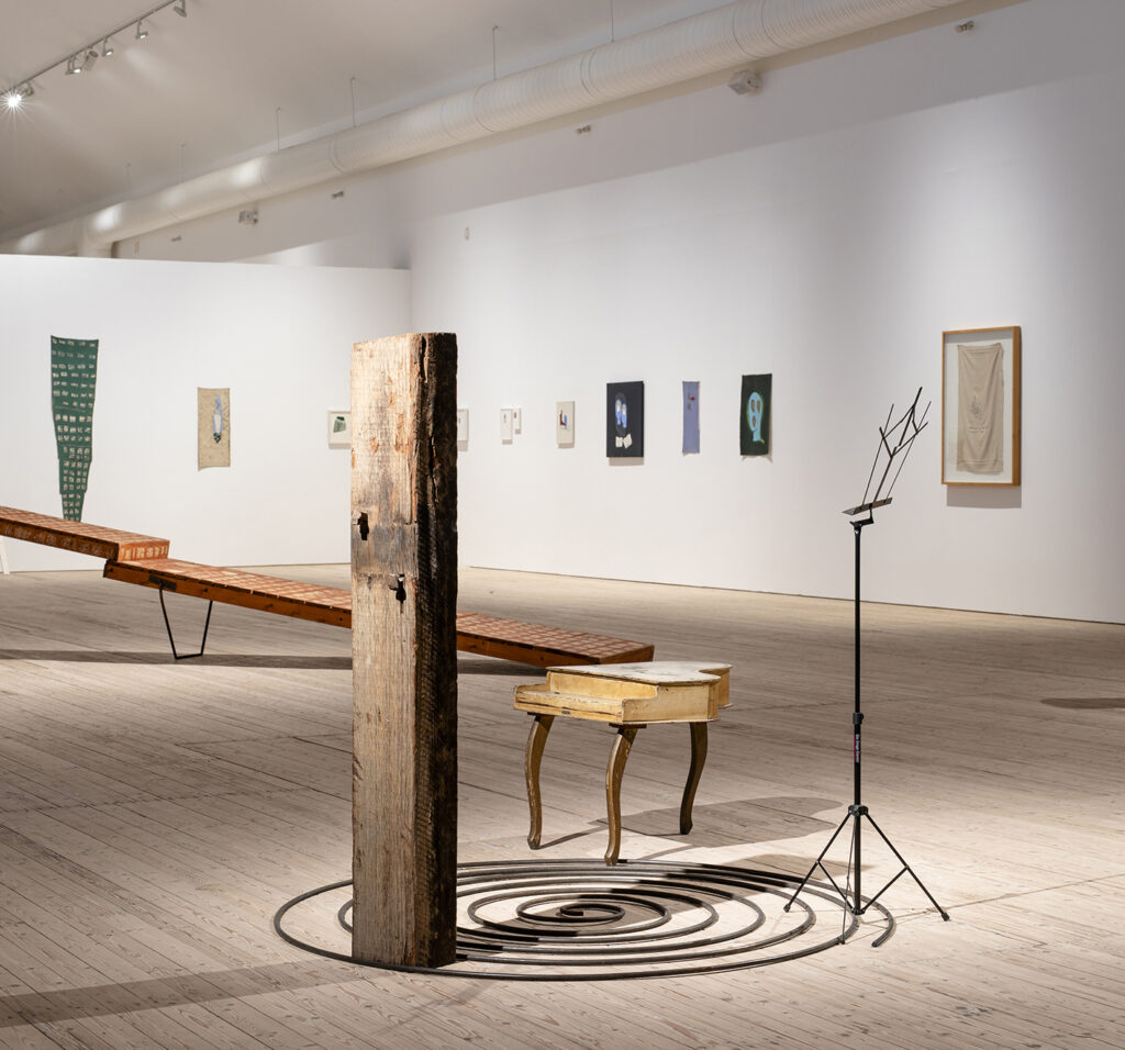 An exhibition view with several paintings in the background and a wooden walkway in the foreground, along with a small wooden piano.