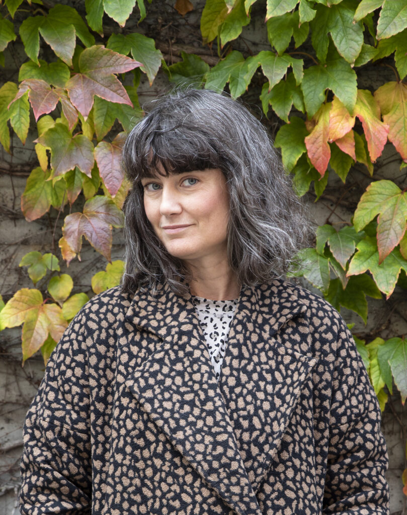 A portrait of Ann Böttcher in front of a wall covered with autumn-colored wild ivy. She is dressed in a leopard-print jacket and is smiling gently.