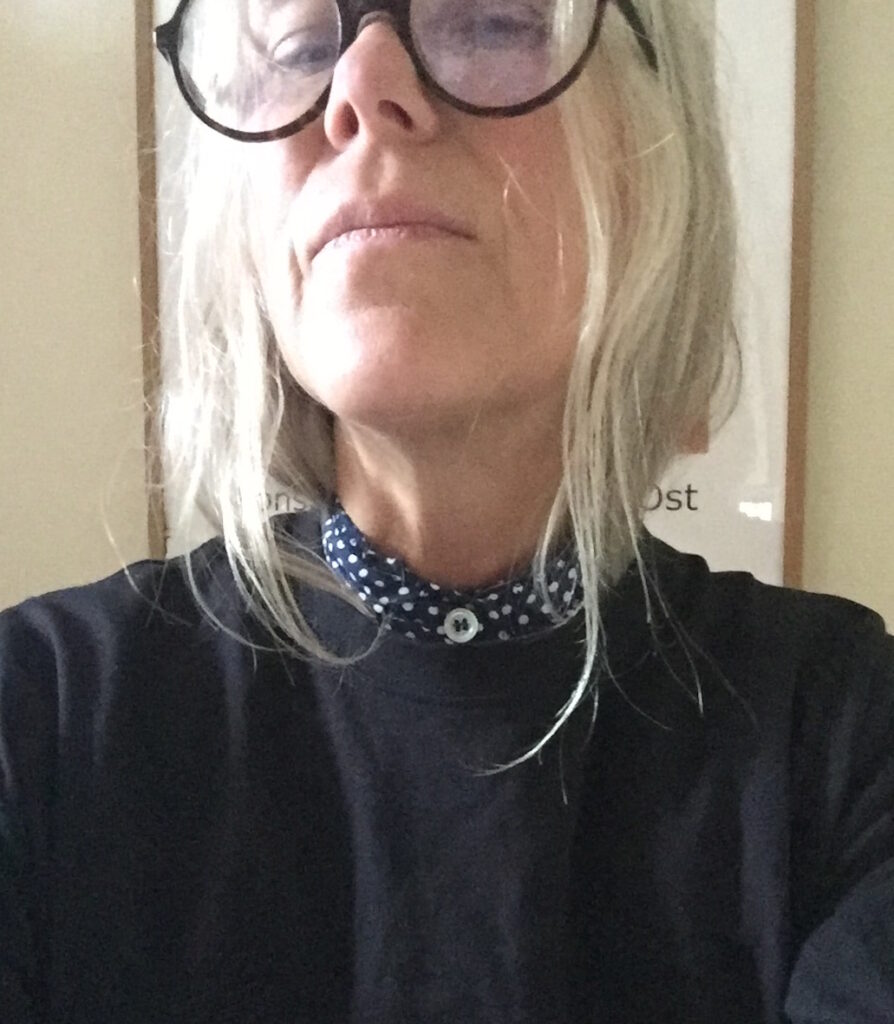 Selfie på kvinna i ljust hår, runda stora glasögon och svart tröja med en prickig skjorta som sticker upp