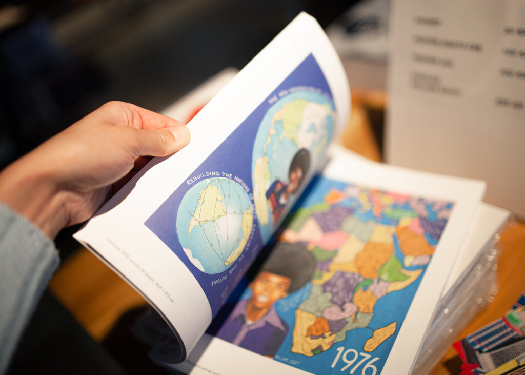 Fotografi av hand som bläddrar i en bok. På sidorna syns färgglada självporträtt av konstnären William Scott tillsammans med jordglobar och kartor