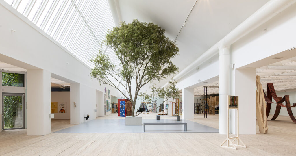 Konsthallen under utställning med olika utplacerade verk i hallen, och i mitten av rummer syns ett stor växande träd