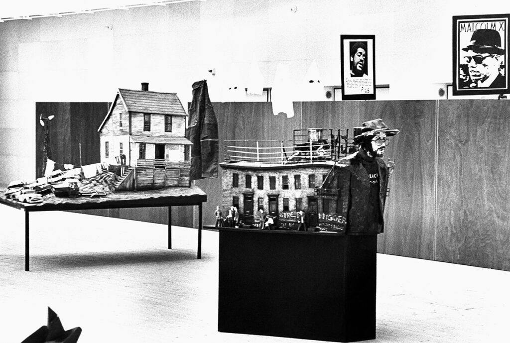 Två miniatyr-hus står uppställda på var sin piedestal i konsthallen. Bakom syns en träpanel och två affischer.