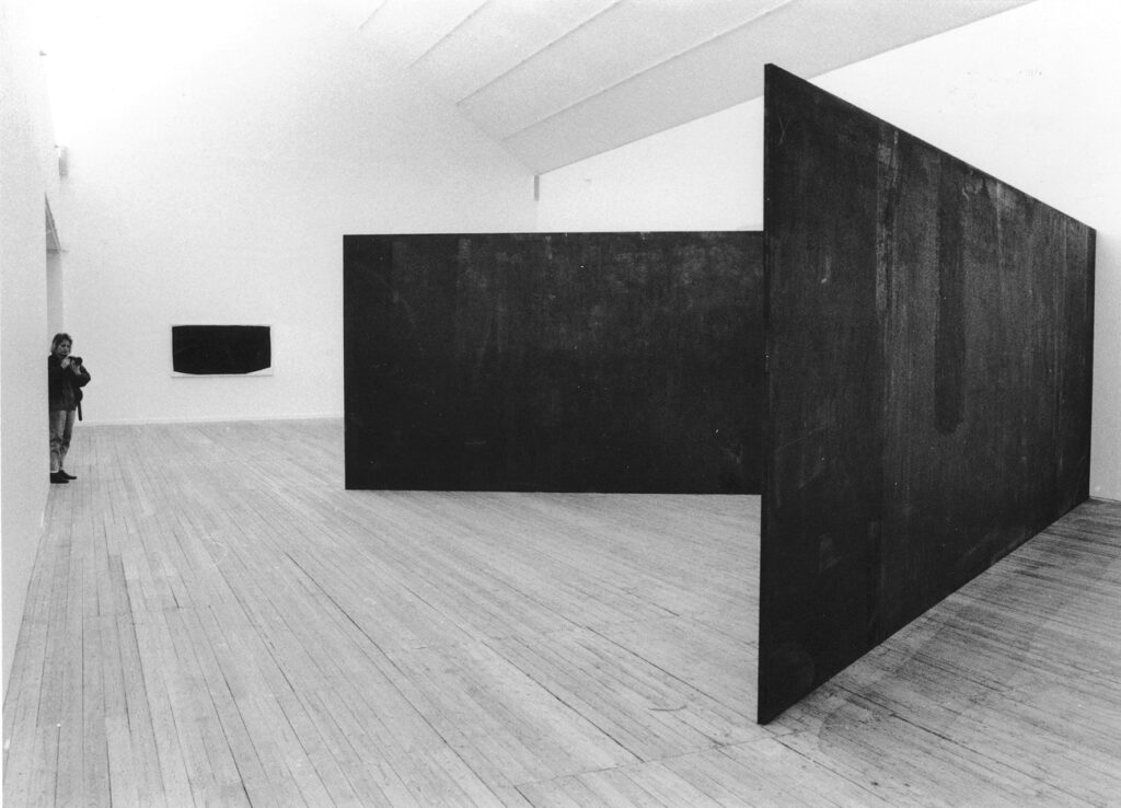 Ett svartvitt fotografi från ett stort rum med vita väggar och ljust trägolv. På väggen längst bort syns en mörk rektangel, till vänster i bild, en bit bak, står en person med en kamera och till höger i bild, närmast där fotografiet är taget, står två stora mörka plattor i en V-formation.