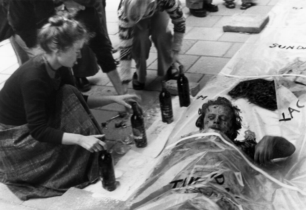 Del ur ett performance. En man försöker ta sig ut ur en stor plastpåse som ligger på gatan. Bredvid honom hukar sig en kvinna som håller lätt i två av fyra vinflaskor som står på rad.