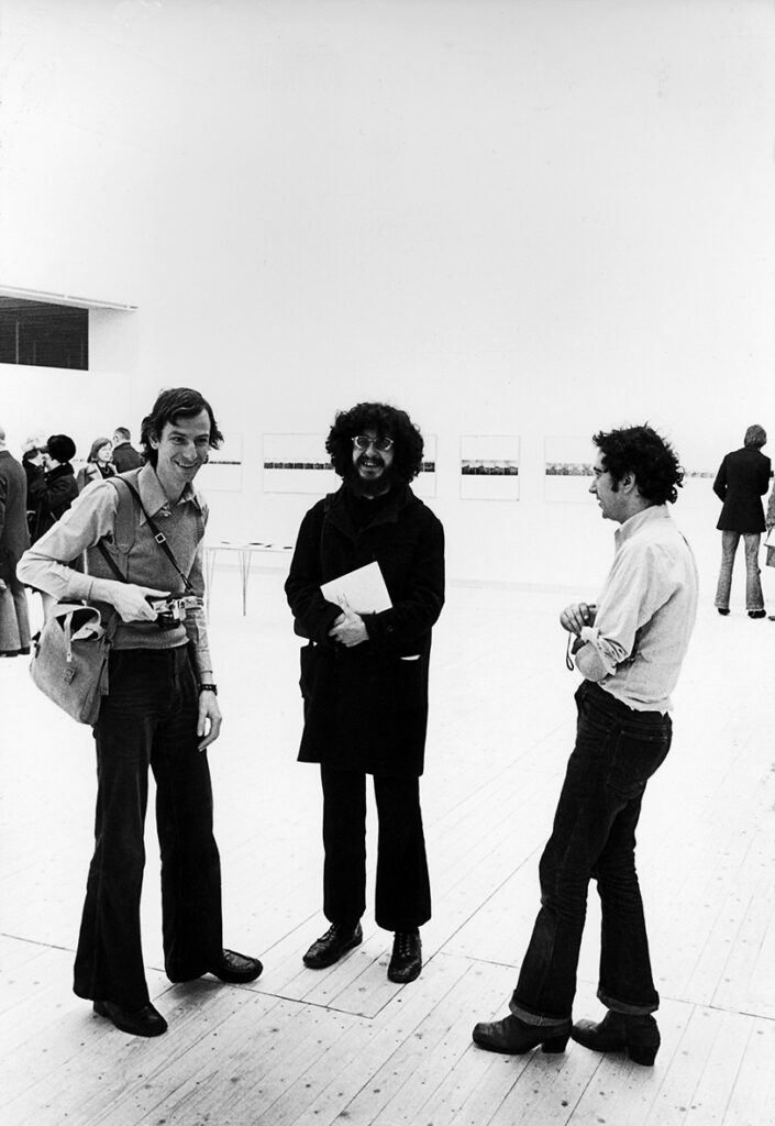 Svartvit bild av tre män som står tillsammans och skrattar. Året är 1975.