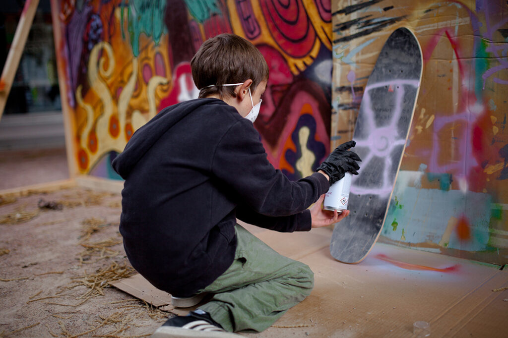 En liten pojke i svart hoodie spraymålar en skateboard.