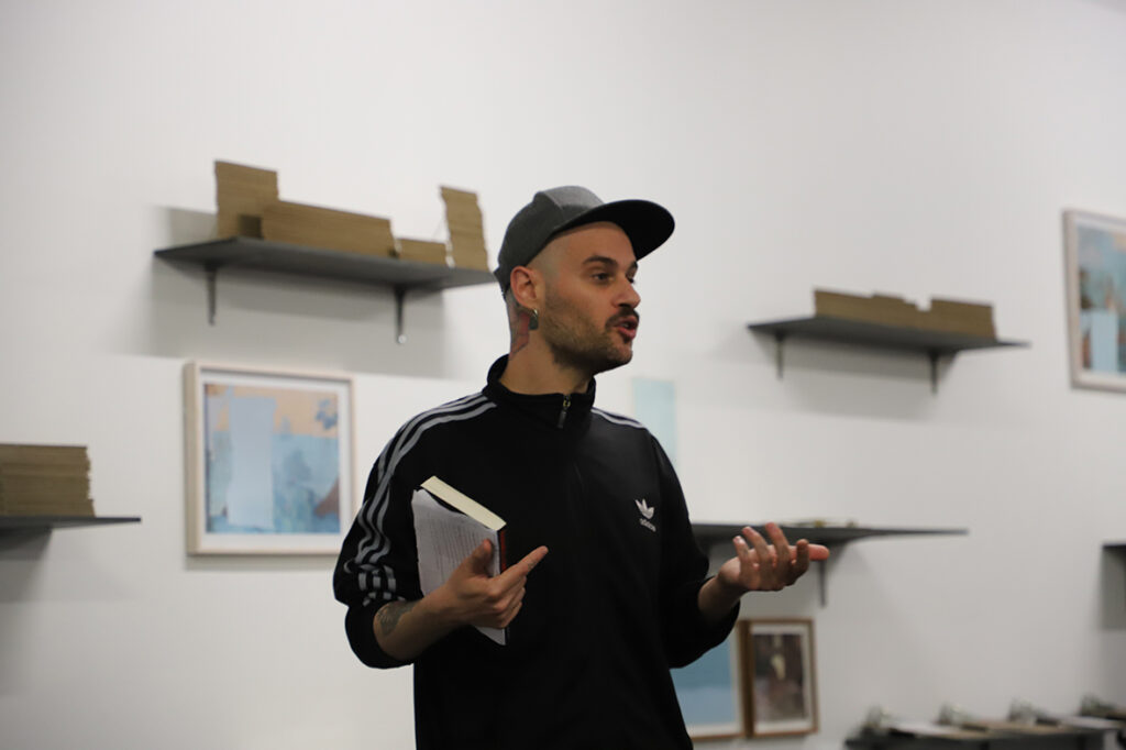 Portrait of Jari Malta talking in front of a fund of wall shelves and holding a book